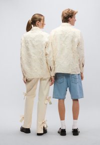 Δύο άτομα φορούν υφασμάτινα μπεζ σακάκια με φθαρμένες άκρες. Το ένα φοράει μπεζ παντελόνια με φιόγκους, το άλλο τζιν σορτς. Και οι δύο έχουν μαύρα παπούτσια.