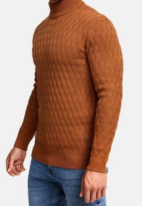Homme portant un pull à col roulé en maille torsadée couleur rouille et un jean bleu, debout avec les bras détendus le long du corps.