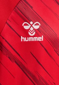 Czerwona sportowa koszulka z teksturowaną tkaniną i czarnymi ukośnymi paskami. Na przodzie znajduje się logo "hummel" w białym kolorze. Krótkie rękawy.