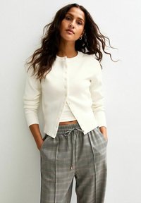 Krämfärgad ribbad cardigan med rund halsringning och knäppning, kombinerad med grårutig byxa med dragsko och sidofickor.