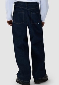 Persona che indossa un paio di jeans larghi blu scuro con tasche posteriori visibili e cuciture a contrasto, abbinati a scarpe nere e a una maglietta bianca a maniche lunghe.