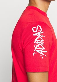 Osoba v červeném tričku s krátkým rukávem, na kterém je vertikálně tištěný bílý text ve stylu graffiti "ADIDAS" na rukávu.