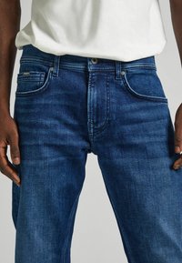 Jeans de denim azul oscuro con un diseño clásico de cinco bolsillos, costuras visibles y un cierre de botón metálico en la cintura.