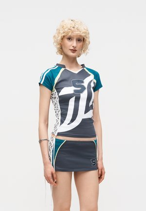 Νεαρό άτομο με ξανθά σγουρά μαλλιά που φοράει αθλητική γκρι, πετρόλ και λευκή στολή με κορδόνια στα πλάγια και το γράμμα "S" πάνω στο μπλουζάκι και τη φούστα.