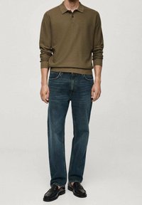 Khaki polo paita, joka on rennoissa mitoissa, yhdistettynä tummansinisii suoriin farkkuihin. Malli käyttää mustia siistipolkuja, seisten neutraalilla taustalla.