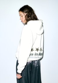 Fehér kapucnis pulóver, amelyen csillagok és szöveg grafikai mintája található a háton. A modell fényes fekete szoknyát és gyűrűket visel.