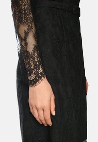 Schwarzes Spitzenkleid mit kunstvollen Spitzenärmeln, strukturiertem Stoff und tailliertem Design. Die Ärmelenden weisen eine fransige Detailverarbeitung auf.