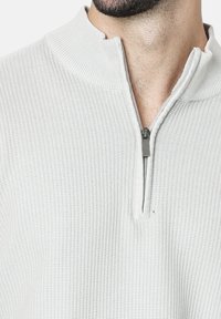 Suéter gris claro con cuello con cremallera, que presenta un patrón texturizado y acanalado, y un sutil brillo en la tela.