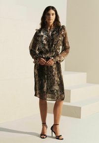 Robe noire et brune en tissu transparent avec un imprimé serpent, épaules volantes, taille cintrée et manches longues, accompagnée de sandales à talons hauts noires.