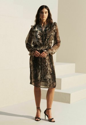 Robe noire et brune en tissu transparent avec un imprimé serpent, épaules volantes, taille cintrée et manches longues, accompagnée de sandales à talons hauts noires.