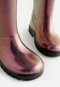 Iridiscenční gumové deštní boty s lesklým fialovým a růžovým gradientem, texturovaným povrchem a černými vroubkovanými podrážkami pro lepší trakci.