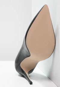 Chaussure à talon haut en cuir noir, avec un bout pointu, une texture lisse et un talon aiguille fin ; la semelle présente des rainures verticales.