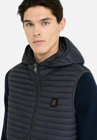 Veste matelassée bleu marine avec capuche amovible, caractérisée par une texture matelassée horizontale, une fermeture éclair à l'avant et un écusson logo sur la poitrine.