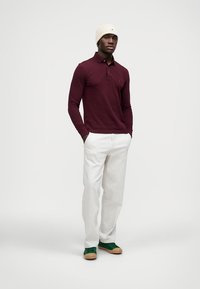 Camisola polo de manga longa em bordô, calças brancas, sapatilhas verdes e um gorro creme. Apresenta um logotipo discreto na polo.