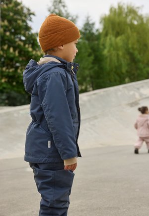 Marineblaue wasserdichte Jacke mit Kapuze und beigen Akzenten, kombiniert mit passenden Hosen. Das Kind trägt eine orange gestrickte Mütze. Außenszene.