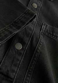 Melnā denima auduma ar gludu tekstūru, kurai ir metāla pogas un kontrastējoša dūka. Tuva skata uz apkakles un kabatas detaļām.