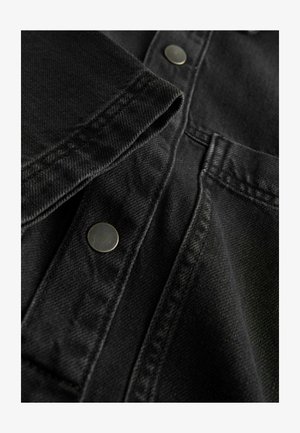 Juodas džinsinis audinys su lygiu paviršiumi, apipavidalintas metaliniais mygtukais ir kontrastingais siūlais. Artimas žvilgsnis į apykaklės ir kišenių detales.