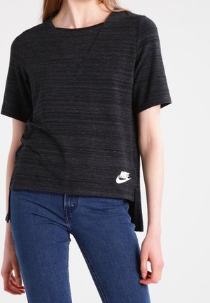 Kobieta ubrana w czarną koszulkę Nike z krótkim rękawem z białym logo na dolnej krawędzi oraz niebieskie dżinsy, stojąca na jednolitym białym tle.