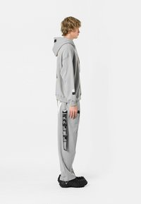 Sudadera gris claro con cordón y estampado gráfico, combinada con pantalones de chándal a juego que presentan detalles en negro y un corte holgado.