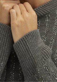 Pull en tricot gris avec des poignets côtelés, comportant des fils métalliques tissés dans le tissu. Gros plan sur des mains tenant le col.
