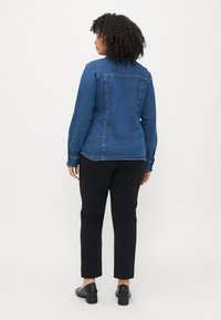 Jeansowa kurtka w średnim niebieskim kolorze z tylnym szwem, długimi rękawami i mankietami na guziki, w połączeniu z czarnymi spodniami typu slim fit i czarnymi butami na obcasie.