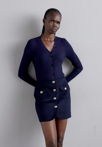 Navy ribbed cardigan with a v-neck and six buttons, paired with a matching navy skirt featuring side pockets and gold button accents.