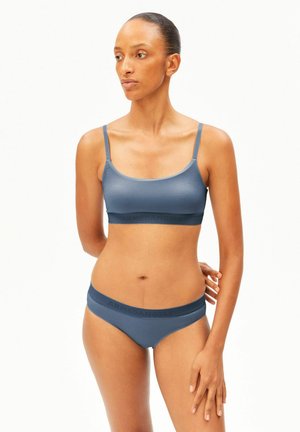 Femme portant un ensemble assorti de soutien-gorge de sport bleu et de sous-vêtements, debout avec une main derrière le dos et regardant sur le côté.