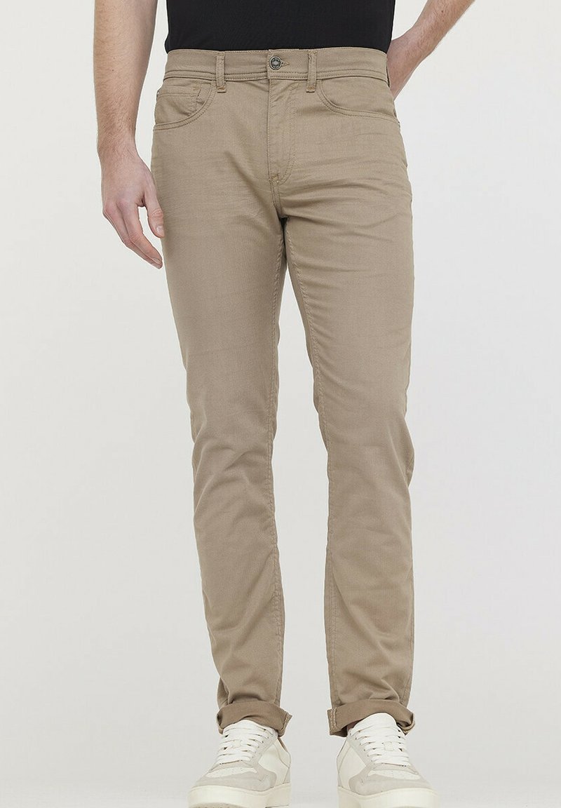 Pantalons slim beige en coton robuste, dotés d'une fermeture éclair, de cinq poches et d'un design de revers, associés à des baskets blanches.