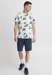 Camisa de manga corta en color crema con estampado floral azul y púrpura, combinada con pantalones cortos cargo en color azul marino y zapatillas deportivas negras con calcetines blancos.