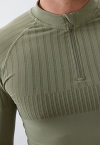 Zöld hosszú ujjú felső magas gallérral és részleges cipzárral, függőleges bordázott textúrával, sima anyaggal és slim fit kialakítással.