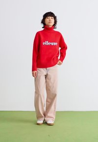 Sarkans adīts turtlenecks ar balto "ellesse" logo, apvienots ar gaiši bežām plašajām biksēm un baltām kedām uz zaļā grīdas.