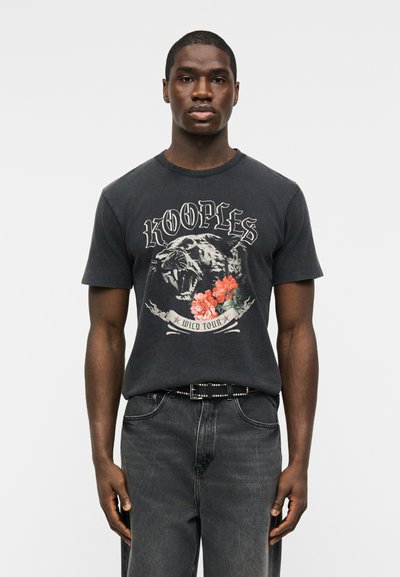 Άνδρας που φοράει μαύρο γραφικό T-shirt με αγριεμένη πάνθηρα, κόκκινα λουλούδια και τις λέξεις «The Kooples Wild Tour», σε συνδυασμό με ξεβαμμένο μαύρο τζιν και ζώνη με καρφιά.