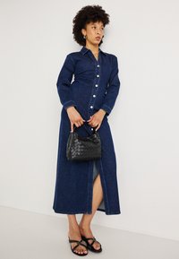 Φόρεμα τζιν με μακριά μανίκια και κουμπιά μπροστά, σκούρο μπλε χρώμα, με άνοιγμα στο πλάι. Μαύρη πλεκτή τσάντα χειρός και μαύρες σανδάλια με διακοσμητική αγκράφα.