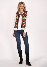 Schwarze bestickte Weste mit floralen Mustern, getragen über einer weißen Bluse, kombiniert mit blauen Jeans und schwarzen westernhaften Stiefeln.