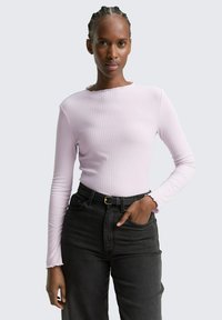 Vrouw met gevlochten haar in een lichtroze ribgebreide langemouwen top, gestopt in een hoge taille zwarte jeans met een zwarte riem, hand in de zak.