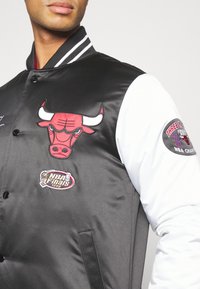 Chaqueta bomber de satén negra con mangas blancas, que presenta un destacado logo rojo de los Bulls y un parche de "NBA Finals 1998", cierre clásico de botones a presión.