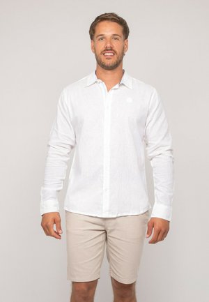 Homme debout portant une chemise blanche à manches longues boutonnée et un short beige arrivant au genou, sur un fond uni.