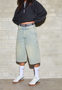 Kort svart jacka, ljusblå denimshorts, vita sneakers med gröna detaljer, vita ankelsockor, med Puma-logotyp på midjebandet.