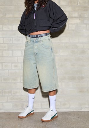 Cropped black jacket, light blue denim shorts, white sneakers with green accents, white ankle socks, featuring Puma logo on the waistband.