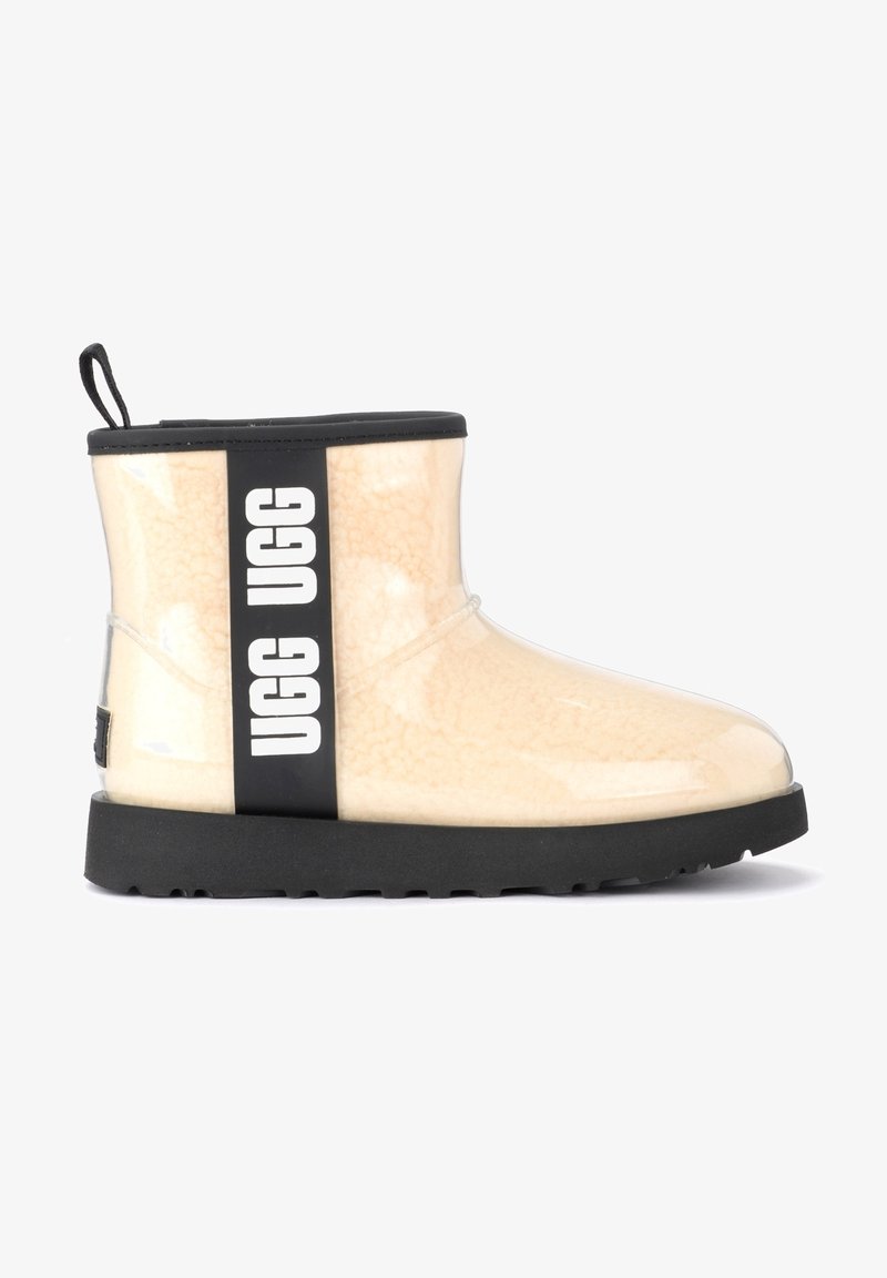 Bottine en mouton beige avec semelle en caoutchouc noire, garniture noire et bande verticale noire avec le logo "UGG" en blanc sur le côté.
