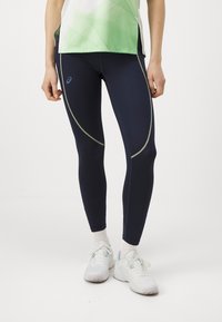 Marineblauwe performance legging met groene stiksels, aansluitend ontwerp en een gladde textuur, gecombineerd met witte sport schoenen en sokken.