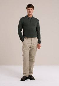 Man in een donkergroene lange mouw polo, beige broek en donkerbruine schoenen, staand met één hand in zijn zak op een effen achtergrond.