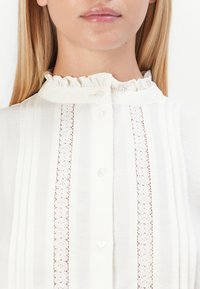 Hvid bluse med flæsekrave, lodrette blondestofpaneler og perle-lignende knapper, båret af en person med langt, blond hår.