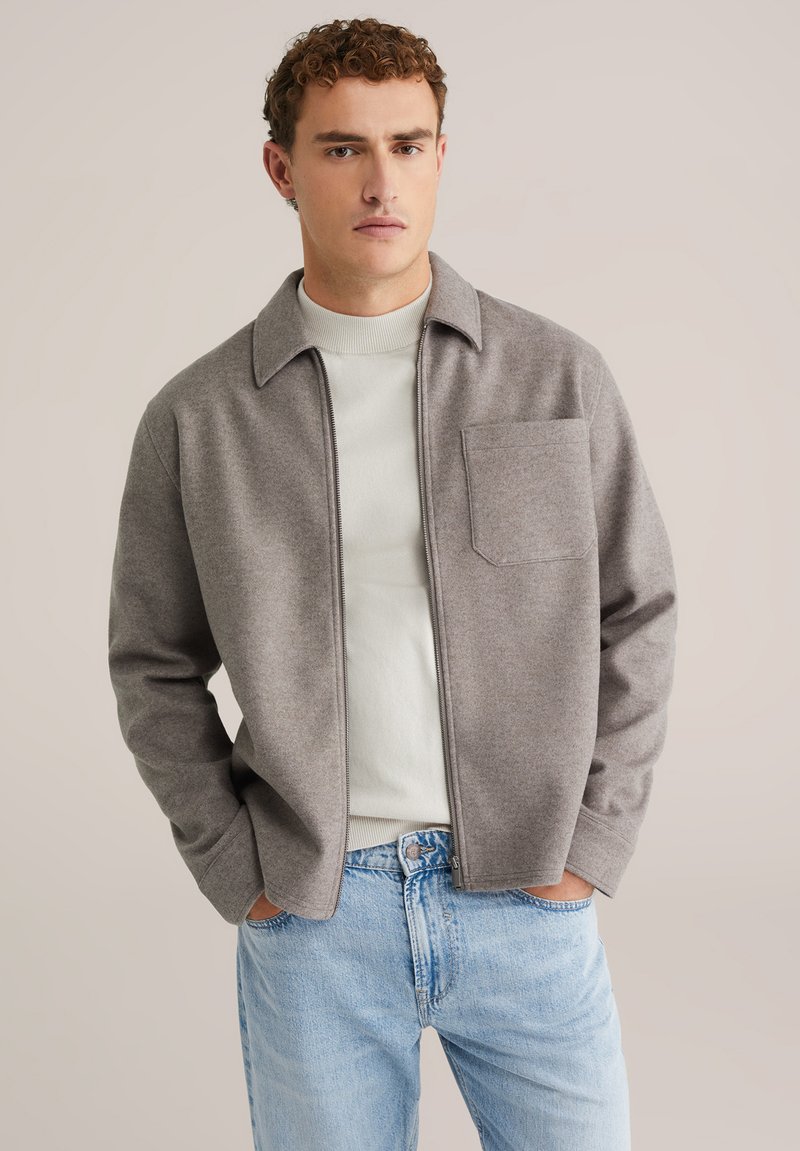 Grijze ritssluitingjack met een puntige kraag, enkele borstzak en gecombineerd met een lichte trui, in combinatie met blauwe jeans. Soepele textuur.