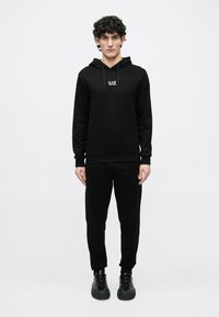 Jauns vīrietis ar cirtainiem matiem, ģērbies melnā EA7 kapucjakā, saskaņotās sporta biksēs un melnās sporta apavos, stāvot pret vienkāršu baltu fonu.