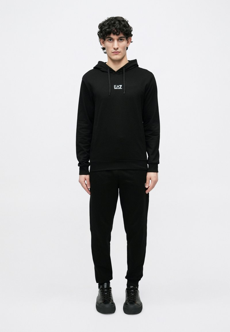 Jauns vīrietis ar cirtainiem matiem, ģērbies melnā EA7 kapucjakā, saskaņotās sporta biksēs un melnās sporta apavos, stāvot pret vienkāršu baltu fonu.