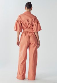 Mono color durazno con cuello tipo camisa, mangas abullonadas y cintura ceñida. Confeccionado en una tela ligera, presenta pantalones de pierna ancha.