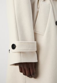 Beige ullfrakk med et strømlinjeformet design, som har en synlig lomme og en mansjett med en svart knappeakzent. Myk tekstur og skreddersydd passform.