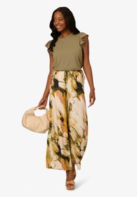 Olivově zelený top s volánovými rukávy, spárovaný s kalhotami s květinovým vzorem ve béžové, žluté a zelené barvě. Drží béžovou kabelku.