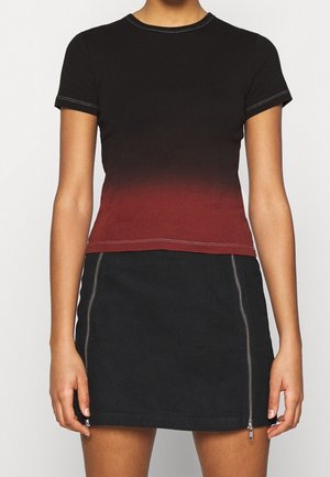 Nuori henkilö pukeutuneena istuvaan mustasta punaiseen liukuvärjättyyn lyhythihaiseen yläosaan ja mustaan minihameeseen, jossa on kaksi pystysuoraa etuvetoketjua.